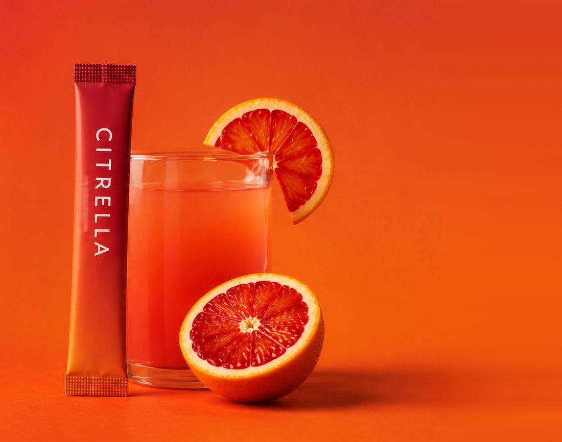 Citrella slim stick standing beside a glass of blood orange drink and two orange halves